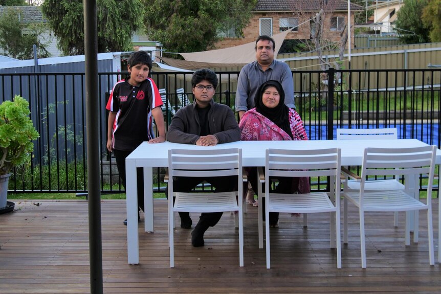 A family, including two boys, a mother and a father sit at an outdoor dining table on a verandah.