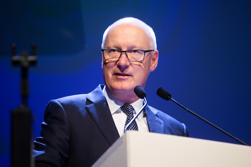 A man with a blue suit and glasses speaks at a corporate lecturn