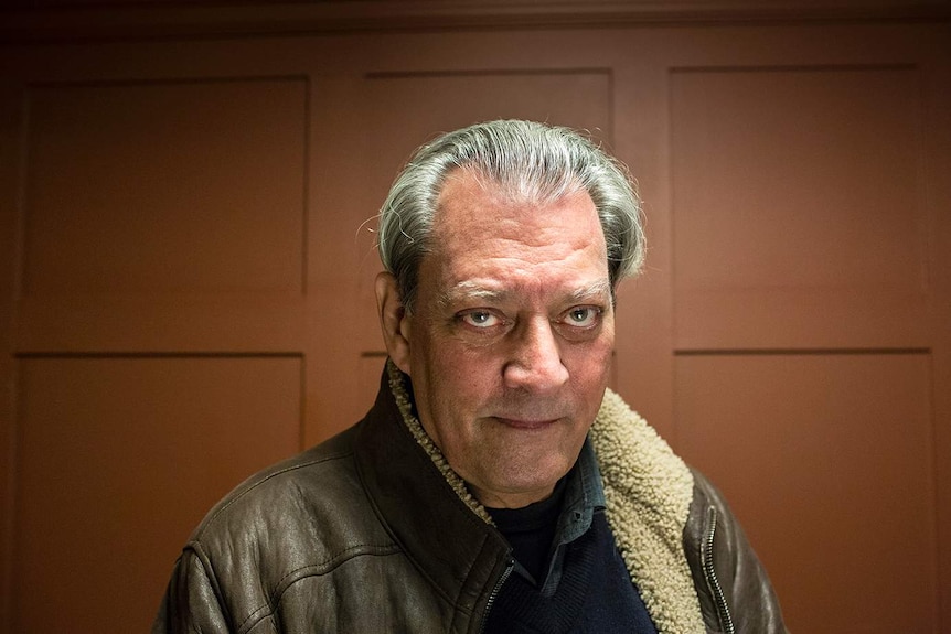 Colour photograph of author Paul Aster standing inside a dimly lit room in front of a brown wall.