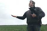 A man holding a dog in a field of crops, holding his hand up looking for rain.