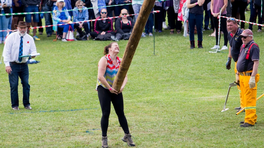 Front on, elevated view of a female competitor tossing a caber as she is watched by onlookers.