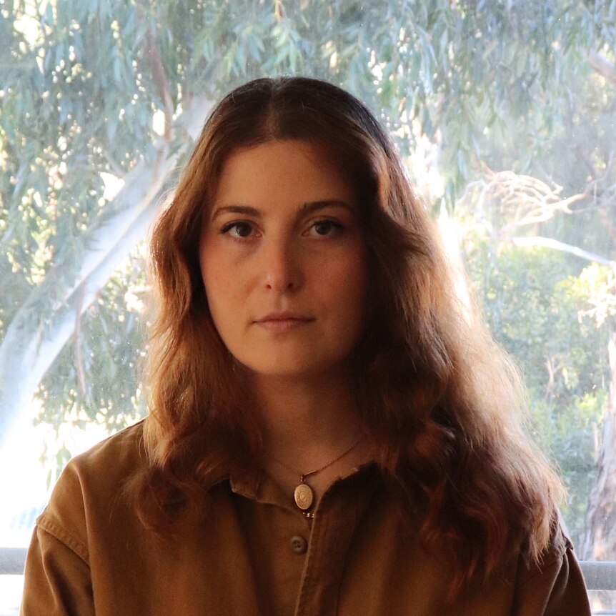 Ella Baxter wearing a brown top with trees in the background through a window.