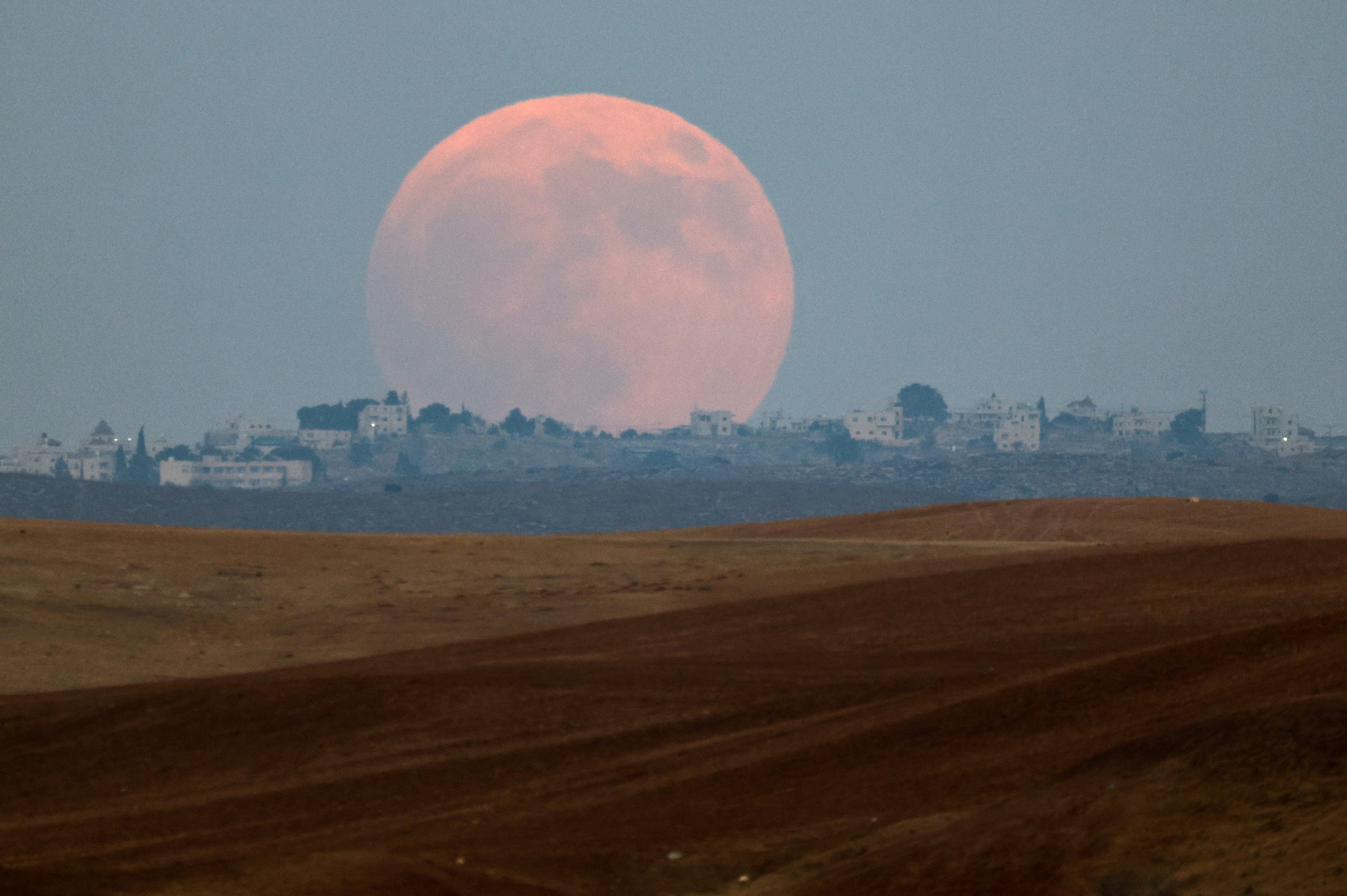 A huge, faint blood moon is seen rising over a minimal landscape, with desert sand in the foreground