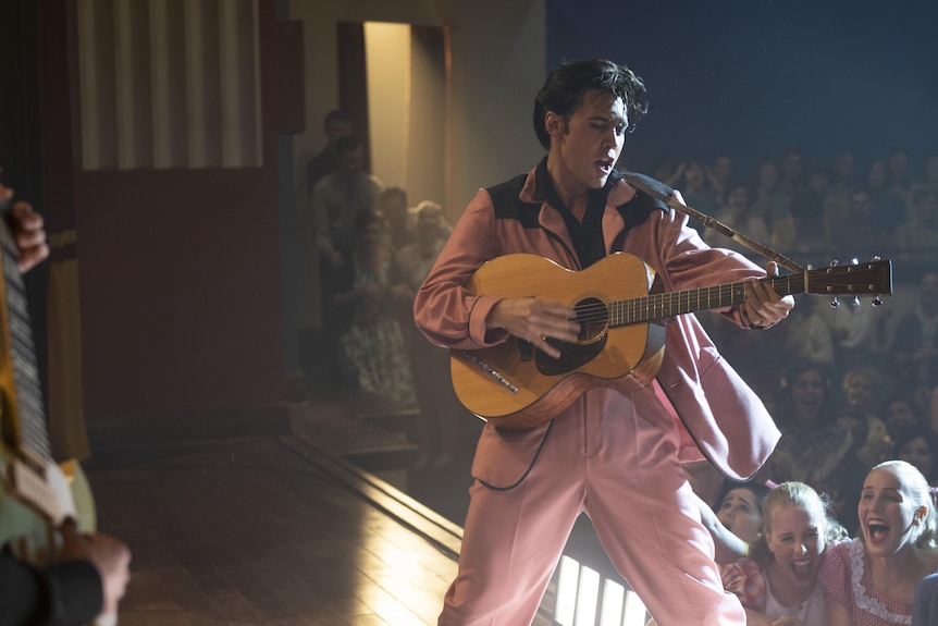 A still image taken from a film about Elvis showing the actor portraying the singer performing in a pink suit with a guitar.