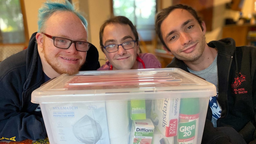 Three young men sitting behind a tub containing supplies for COVID-19 patients