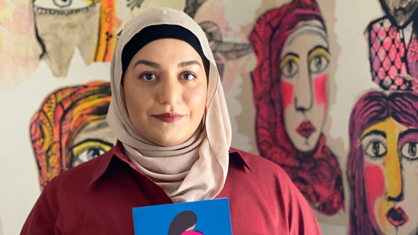 Author Amani Haydar is wearing a blush coloured hijab and holding a copy of her book The Mother Wound