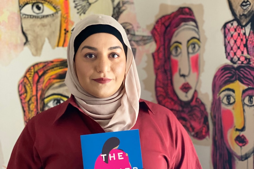 Author Amani Haydar is wearing a blush coloured hijab and holding a copy of her book The Mother Wound