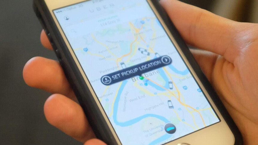 An Uber user holds a mobile phone displaying the app screen with the location set to Brisbane