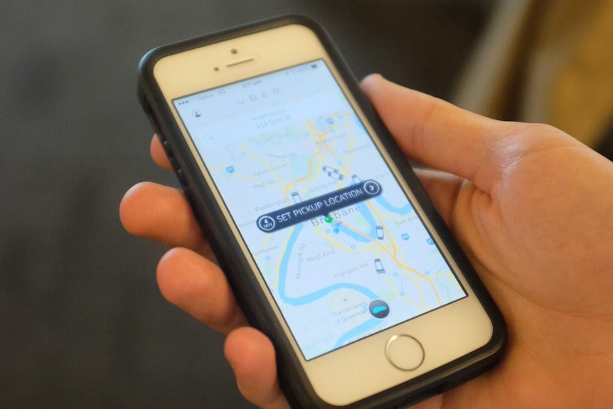 An Uber user holds a mobile phone displaying the app screen with the location set to Brisbane