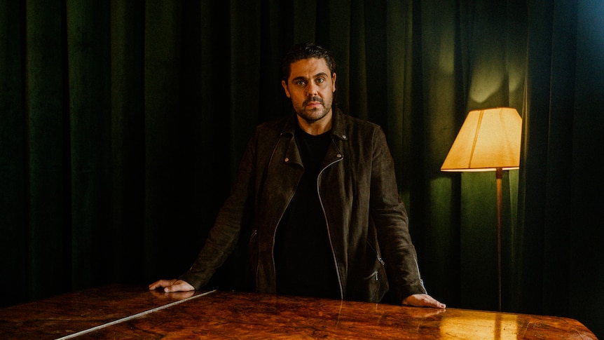 Dan Sultan stands with hands on table looking at the camera against a deep green curtain background