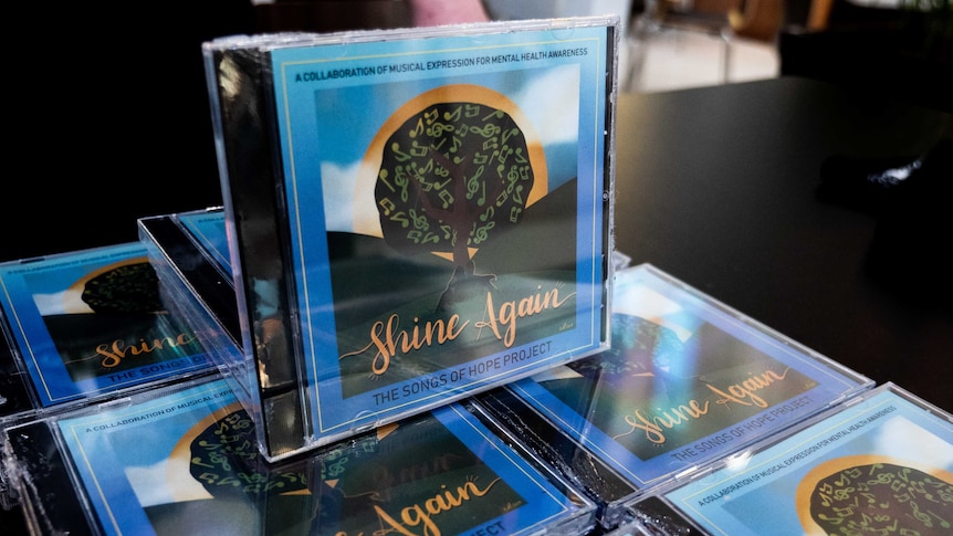 A CD called Shine Again, by The Songs of Hope Project sits on top of a stack of the same CD.