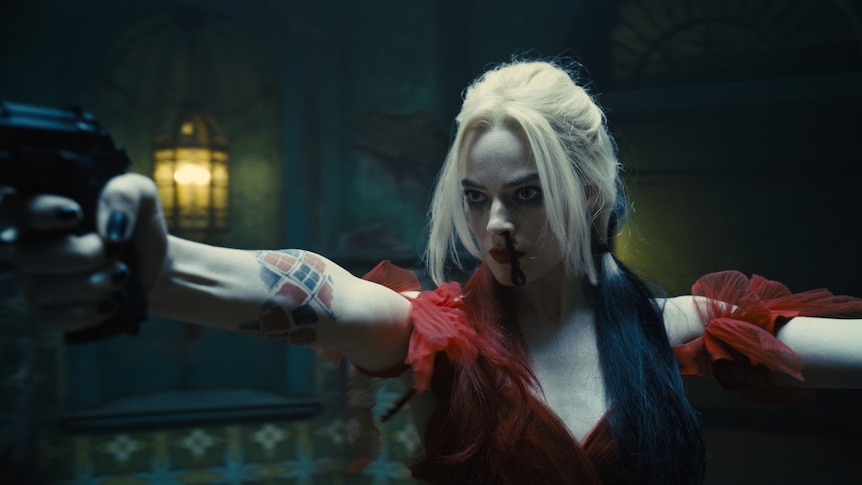 Margot Robbie as Harley Quinn has a bloody nose and is holding a gun in one outstretched hand and wearing a red dress
