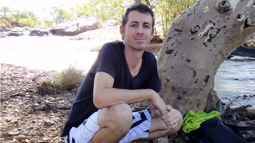 A man in a dark t-shirt and white striped shorts kneels down in a bushland setting.