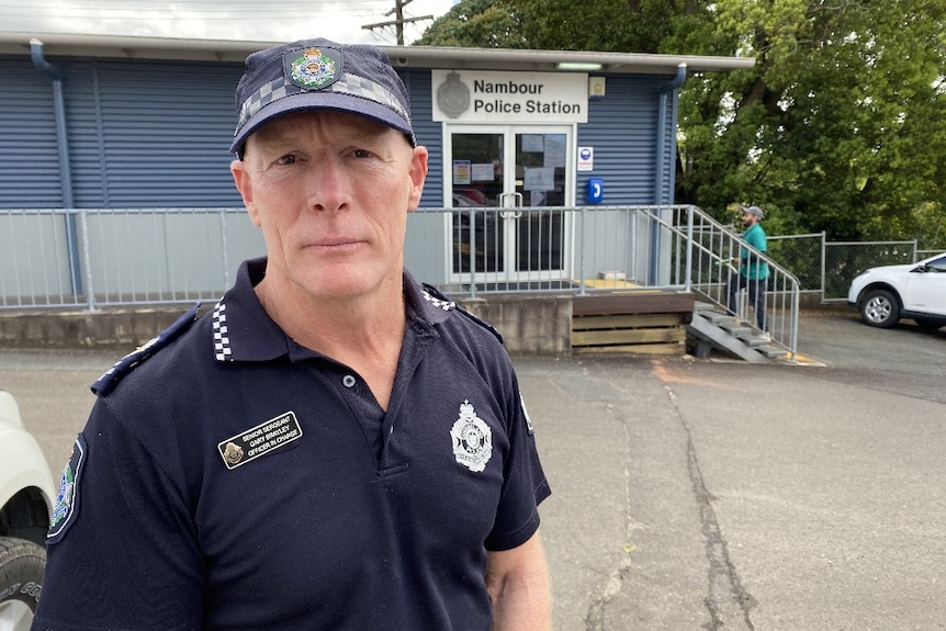 A police officer wearing a hat standing in front of a Nambour Police Station sign.