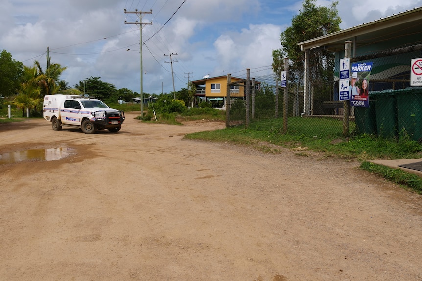 A sandy street in the Torres Strait, with grass and a fenced building and a police divvy van about to park out front