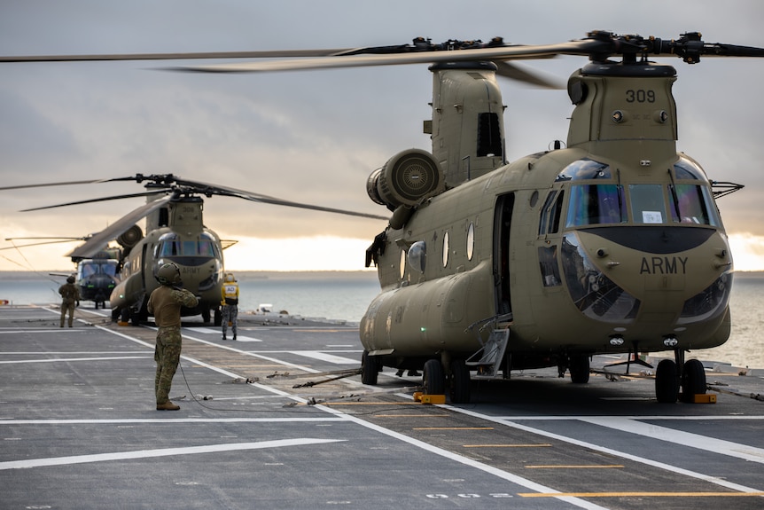Two Chinook helicopters are parked on the deck of HMAS Adelaide with soldiers standing next to them.
