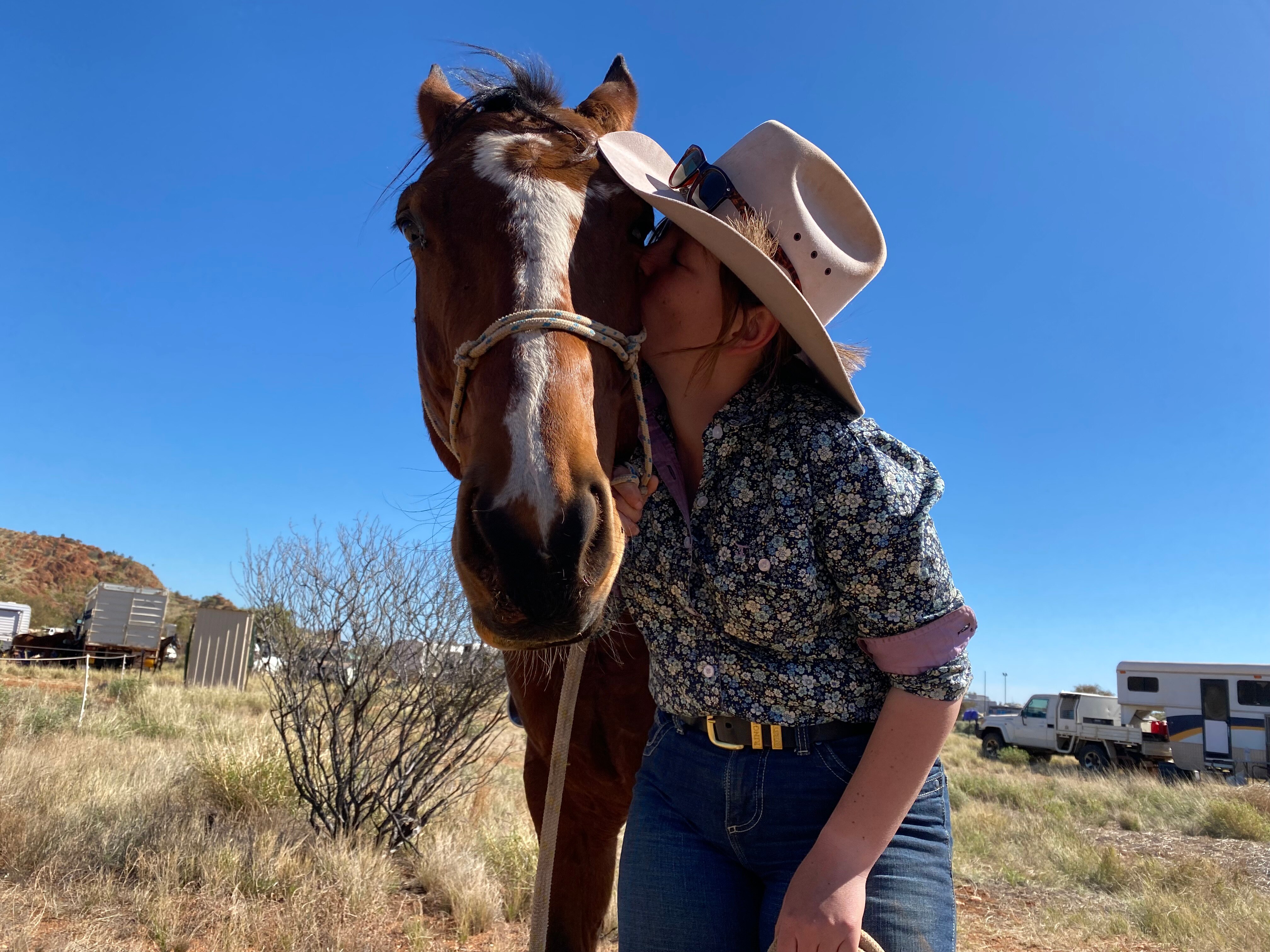 A young woman wearing a floral collared shirt, cowgirl hat and sunglasses kisses the jaw of a bay horse in the outback.