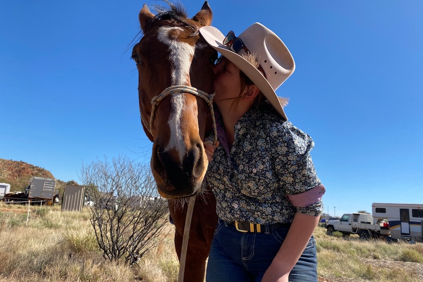 A young woman wearing a floral collared shirt, cowgirl hat and sunglasses kisses the jaw of a bay horse in the outback.