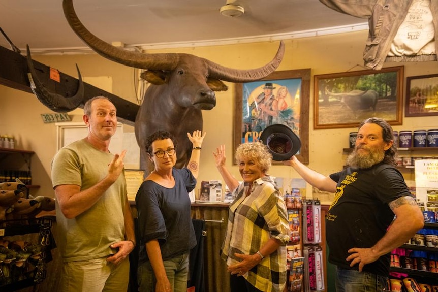 3 men and a woman inside a pub arms outstretched to a giant buffalo head hanging on the wall. 