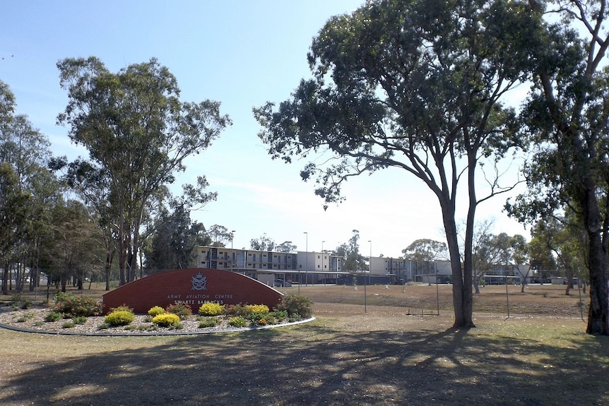 A landscape photo of a brick welcome sign among gum trees with buildings behind it. Between sign and structure is a tall fence