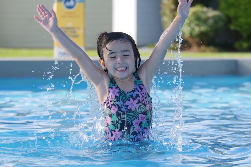 A young girl jumping out of the water, smiling, in a public pool.