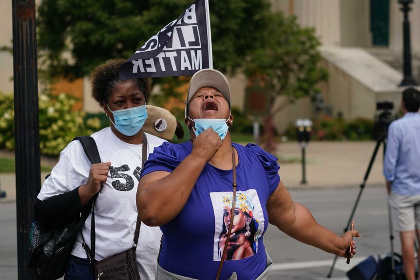 A woman with her head back and mouth open wearing a Breonna Taylor t-short near a Black Lives Matter flag.