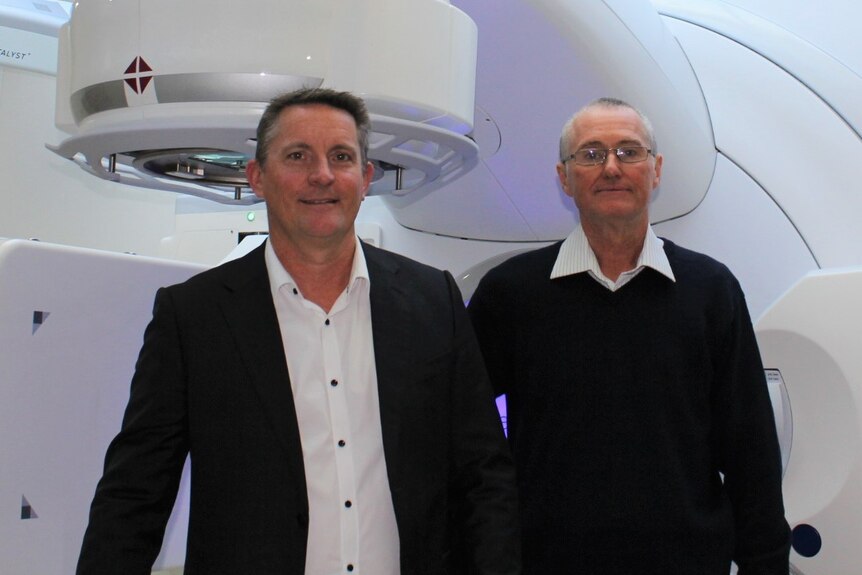 two men standing in front of large white cancer treatment equipment.