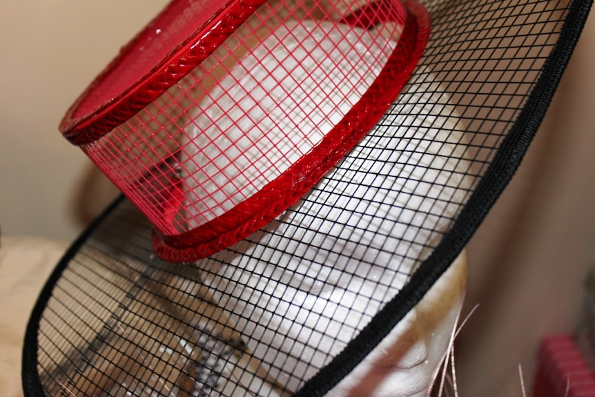 A red and black boater style hat sits on a head mannequin. The hat is made out of gutter mesh.