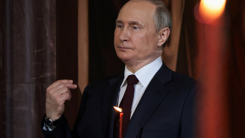 Vladimir Putin looks solemn as he points two fingers at himself in a church.