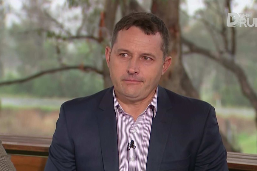 A man with short dark hair, wearing a dark suit, sits in a TV studio.