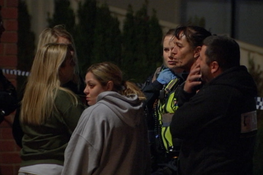 Three young women and a man speak to two uniformed female police officers on the street, with police tape in the background.