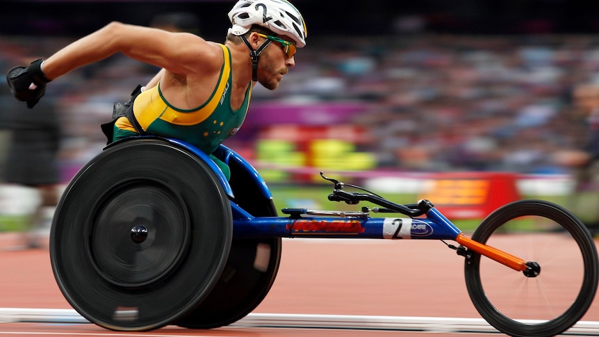 Man in racing wheelchair speeds along athletics tracks in crowded stadium.