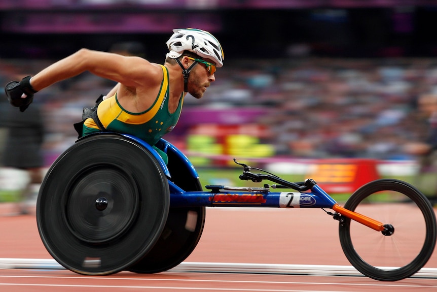 Man in racing wheelchair speeds along athletics tracks in crowded stadium.