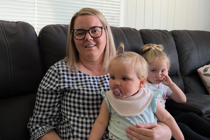 A woman in black glasses and a gingham dress has a baby on her lap and a toddler sitting next to her on a couch