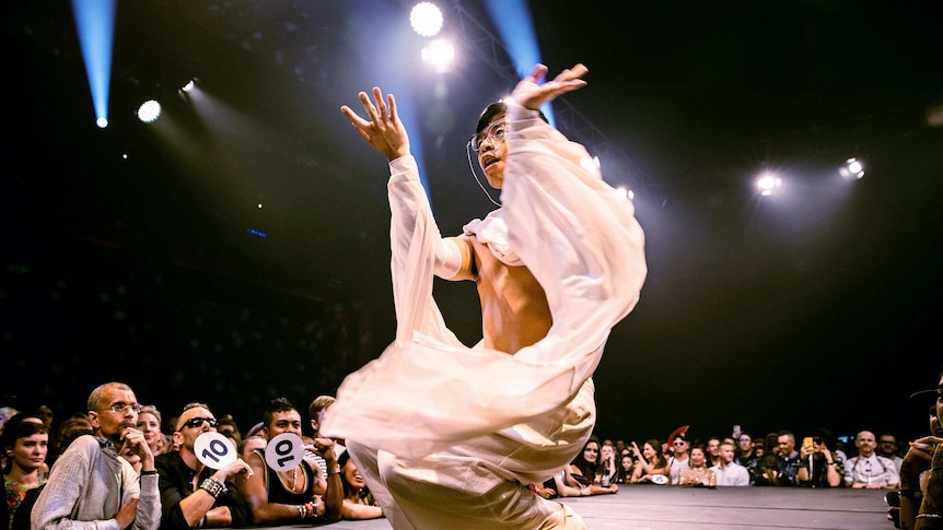A vogue performer in a white, flowing costume crouches on the runway, while audience looks on, at Sissy Ball 2019.