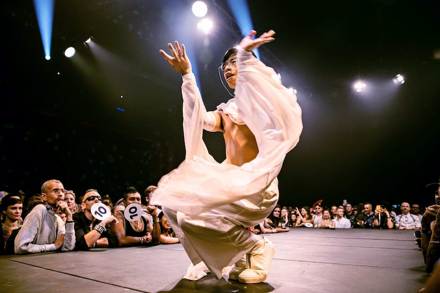 A vogue performer in a white, flowing costume crouches on the runway, while audience looks on, at Sissy Ball 2019.