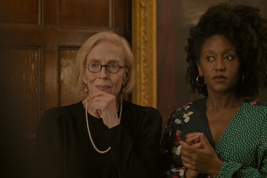 An older white woman in a black jacket and a young black woman in a green top stand in a formal room..