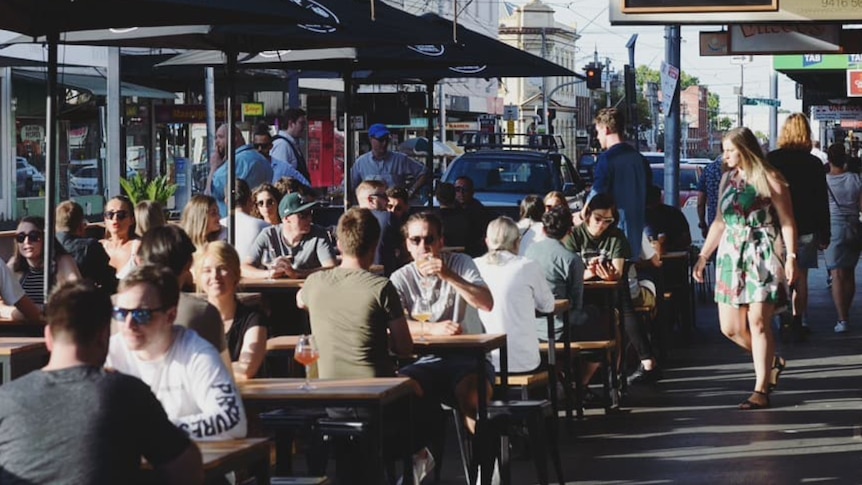 Dozens of people sit out the front of a busy bar.