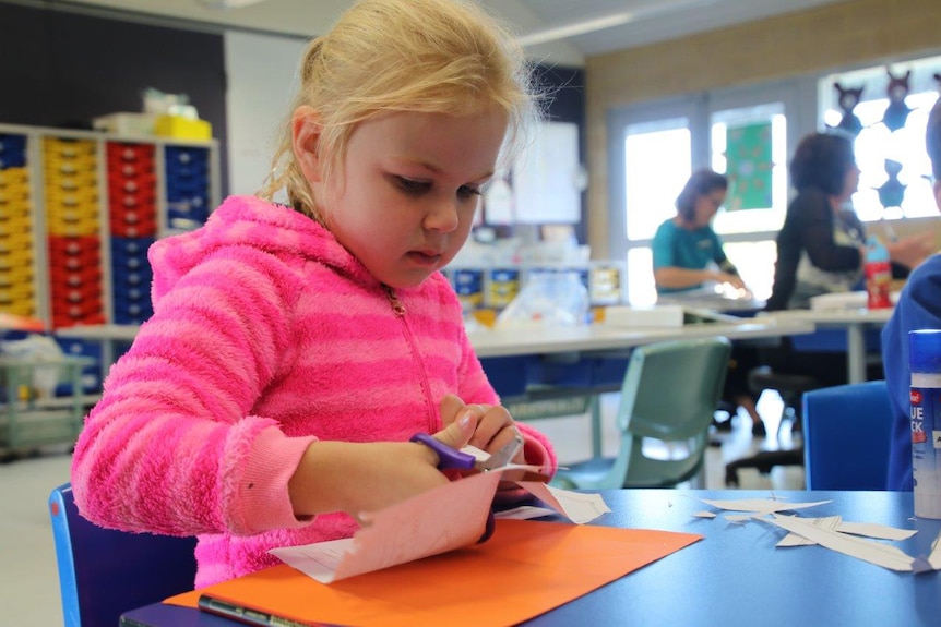 A young girl wearing a pink jumper with blonde hair sits at a table in a classroom cutting paper with scissors.