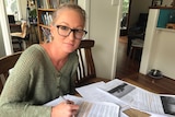 A blonde woman with glasses sits at a desk in her home