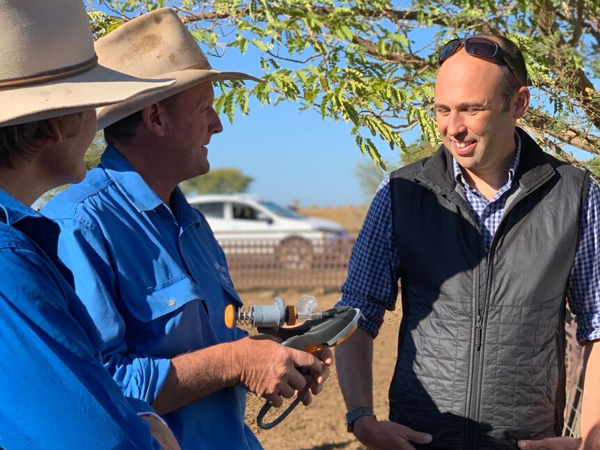 Two farmers in blue shirts and hats hold a device. A man in a puffer vest stands beside them.