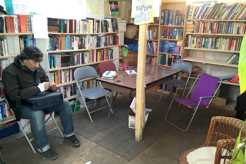 A man sits in the library in the refugee camp, surrounded by a table and chairs and several shelves full of books.