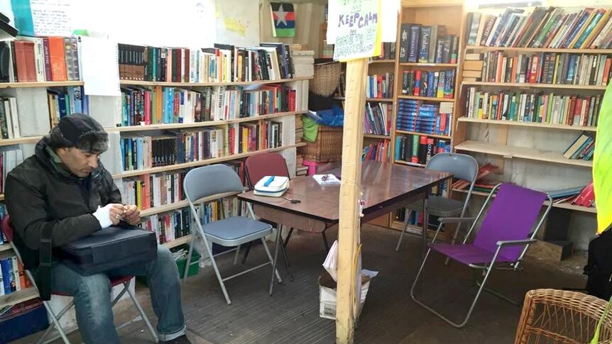 A man sits in the library in the refugee camp, surrounded by a table and chairs and several shelves full of books.