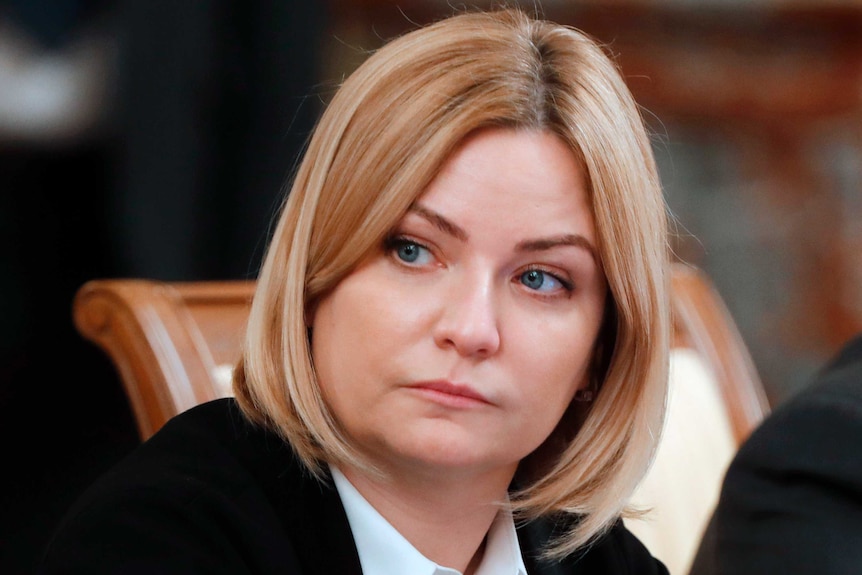 A close up of Ms Lyubimova with blond hair and green eyes glancing off camera during a meeting.
