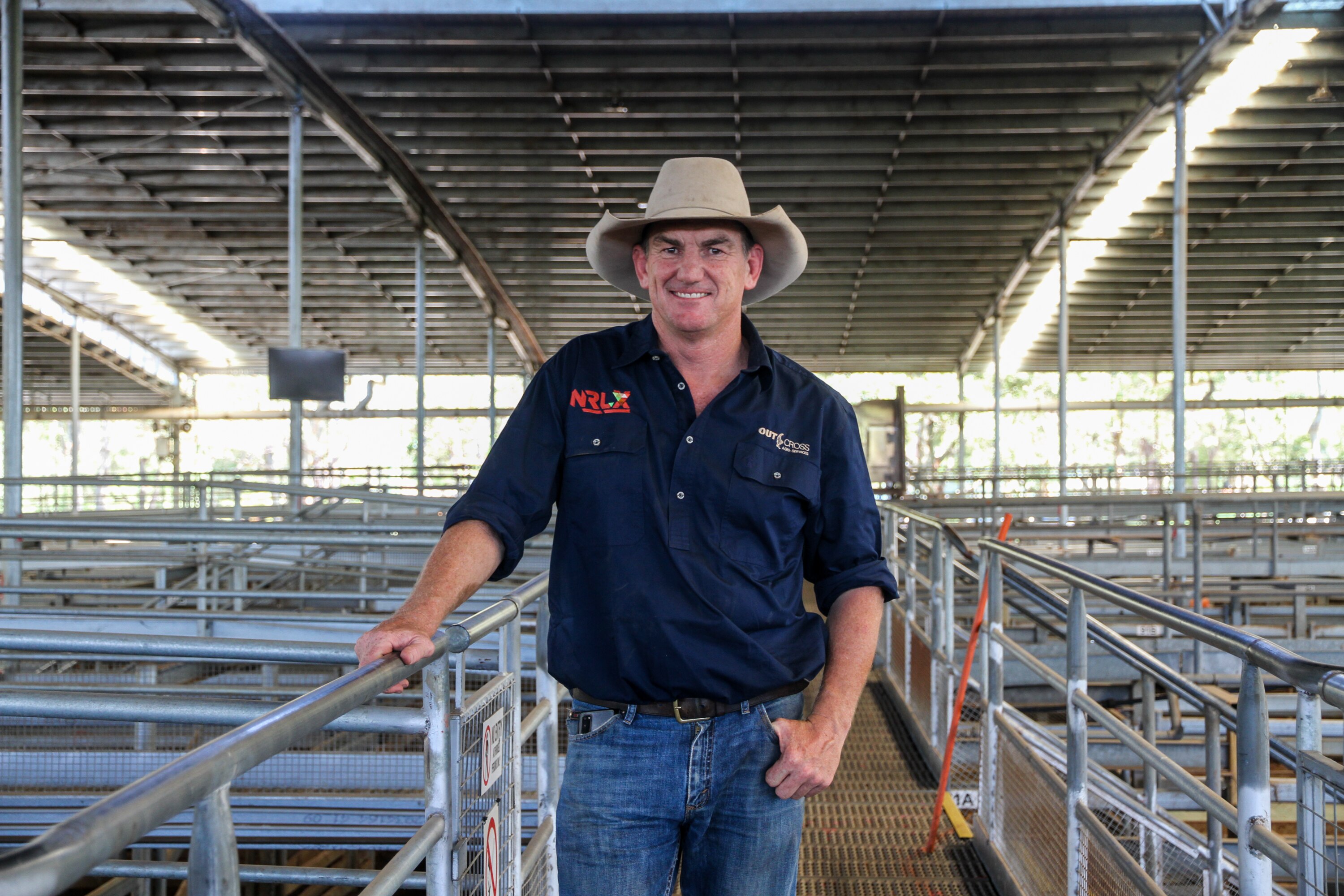 A man in a dark blue shirt and a light broad-rimmed hat holds onto a rail in a large saleyard.