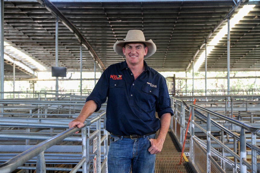 A man in a dark blue shirt and a light broad-rimmed hat holds onto a rail in a large saleyard.