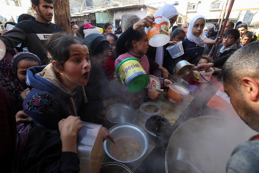 women, children and men crowd around a volunteer cooking holding out buckets and bowls