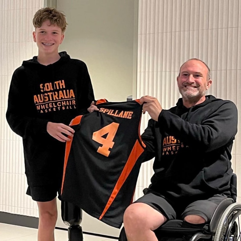 A boy stands and holds a basketball singlet alongside a man in a wheelchair, they smile for the camera.