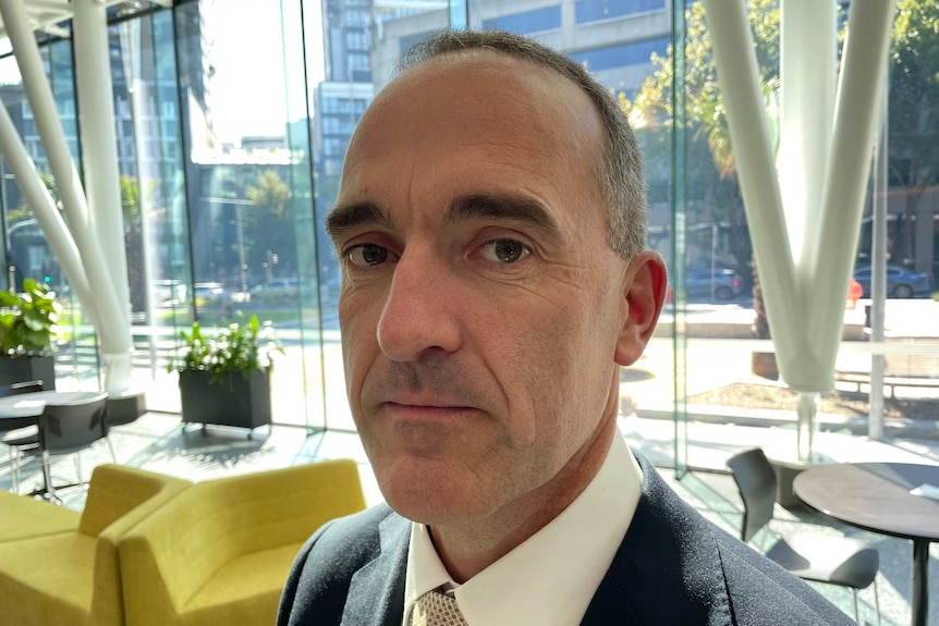 A middle-aged man with short hair stands in an office building, looking at the camera.