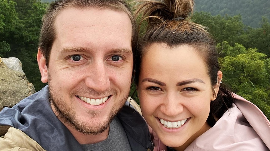 A young man and woman stand grinning while taking a selfie on top of a misty mountain